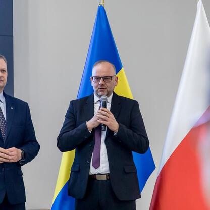 Dwóch mężczyzn w strojach formalnych stoi przed flagami, prawy mówi do mikrofonu, reprezentując Rozwój Lokalny Kierowany przez Społeczność (RLKS) w województwie śląskim. (wygenerowano przez AI)