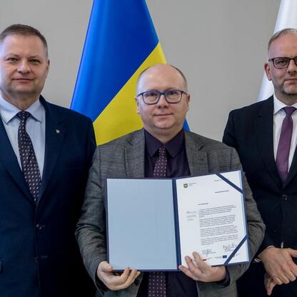Trzech mężczyzn w strojach formalnych stoi przed flagami, reprezentującymi Ukrainę i Polskę, wskazując na dyplomatyczny lub oficjalny kontekst. (wygenerowano przez AI)