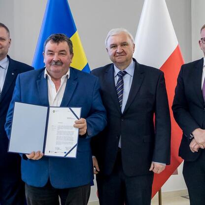 Czterech członków zarządu w ścisłym stroju biznesowym stojących przed flagami polskimi i europejskimi, prezentujących dokumenty, związanych z projektem Rozwój Lokalny Kierowany przez Społeczność. (wygenerowano przez AI)