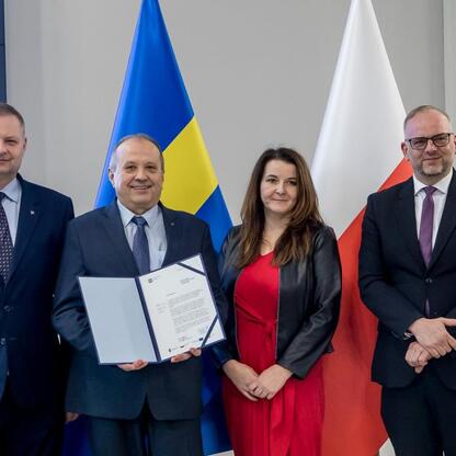 Grupa czterech osób w strojach formalnych, stojących przed flagami UE i Polski, uczestniczących w ceremonii związanych z programem Rozwój Lokalny Kierowany przez Społeczność w Śląskim Województwie. (wygenerowano przez AI)