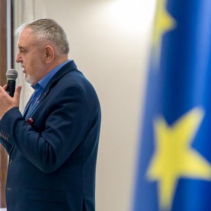 Mężczyzna w ciemnoniebieskim garniturze przemawia do mikrofonu, podczas prezentacji wsparcia dla rodzinnych ogrodów działkowych województwa śląskiego. (wygenerowano przez AI)