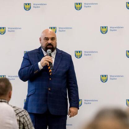 Na scenie stoi wojewoda Śląska, Grzegorz Boski, w niebieskim garniturze, trzymając mikrofon, podczas uroczystości finansowej wsparcia dla rodzinnych ogrodów działkowych. (wygenerowano przez AI)