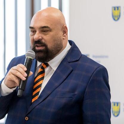 Mężczyzna w niebieskim krawacie z mikrofonem, prezentuje finansowe wsparcie dla rodzinnych ogrodów działkowych w Śląskim. (wygenerowano przez AI)