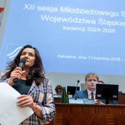 W czasie XII sesji Młodzieżowego Sejmiku Województwa Śląskiego, kobieta przemawia do mikrofonu, trzymając dokument, podczas gdy słuchacze w tle uważnie nasłuchują. (wygenerowano przez AI)