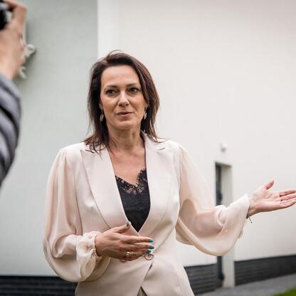 Widoczna kobieta w bluzie wyjaśnia na zewnątrz, przed modernistycznym budynkiem, związana z podpisaniem umowy na budowę nowej sali gimnastycznej w Bażanowicach. (wygenerowano przez AI)