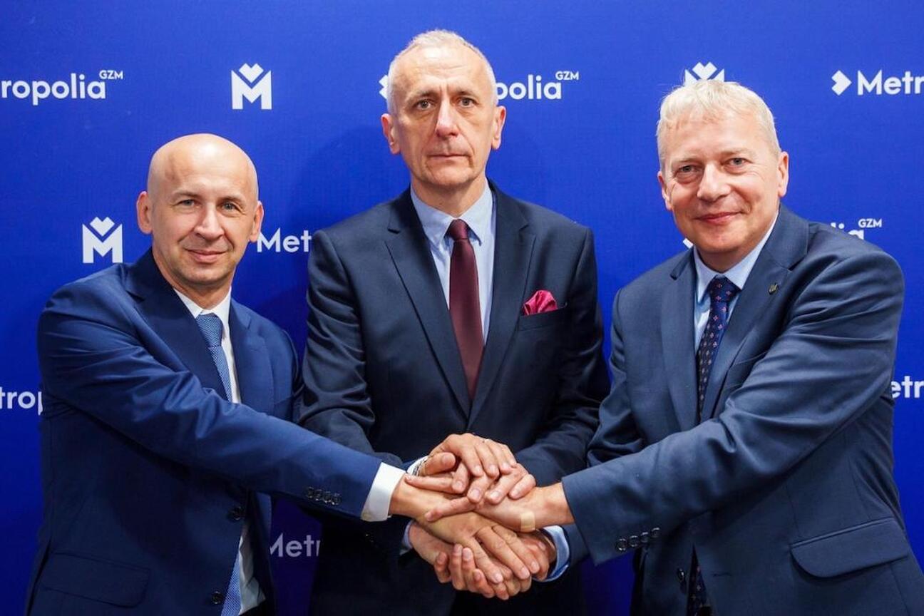 Trzech mężczyzn w strojach formalnych trzyma się za ręce przed logo "Metropolia GZM". (wygenerowano przez AI)