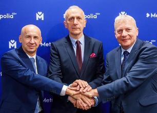 Trzech mężczyzn w strojach formalnych trzyma się za ręce przed logo "Metropolia GZM". (wygenerowano przez AI)