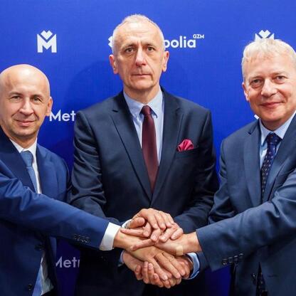 Trzech mężczyzn w strojach formalnych trzyma się za ręce przed logo 