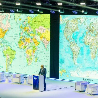 Formalna sesja w Europie Ekonomicznej Kongresie, Katowice, Polska. Mężczyzna przemawia na podium, publiczność siedzi w białych fotelach. Tło z mapami świata. (wygenerowano przez AI)