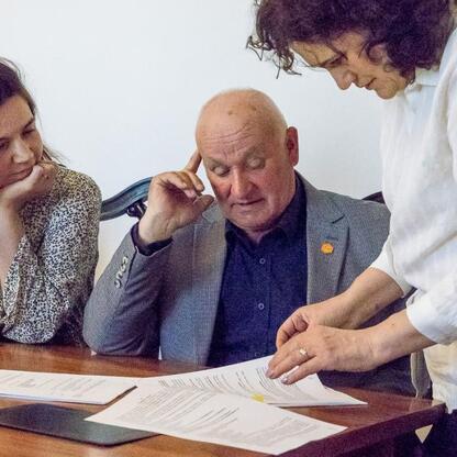 Trzech ludzi w strojach formalnych siedzi przy stole, podpisując dokumenty lub dyskutując w formalnym pomieszczeniu. (wygenerowano przez AI)