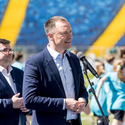Mężczyzna w ciemnym garniturze przemawia do mikrofonu na stadionie Superauto.pl w Sosnowcu, przygotowując się do imprezy sportowej. (wygenerowano przez AI)