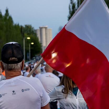 Zdjęcie przedstawia uczestników drugiego Biegu dla Flagi w Tauron Parku Śląskim, ubranych w biało-czerwone koszulki, biegnących lub idących po ścieżce, z jednym z nich trzymającym duży polski flagę. (wygenerowano przez AI)