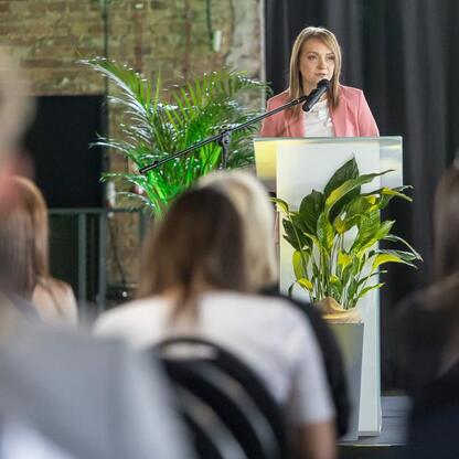 Kobieta w różowej kurtce przemawia na podium w konferencji o rewitalizacji przestrzeni przemysłowych. (wygenerowano przez AI)