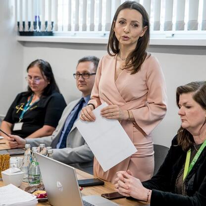 Formalna sesja w konferencyjnym pomieszczeniu, gdzie kobieta w różowej sukience prezentuje Regionalny Plan Adaptacji do zmian klimatu dla Śląska. (wygenerowano przez AI)