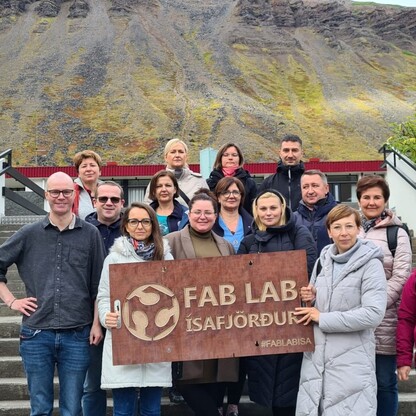 Zdjęcie przedstawia grupę 15 osób, która stoi przed budynkiem i trzyma tablicę z napisem FAB LAB Ísafjörður.