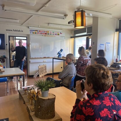 Zdjęcie przedstawia grupę siedzących osób na warsztacie edukacyjnym w FAB-LAB-ie oraz osobę prezentującą przy tablicy.