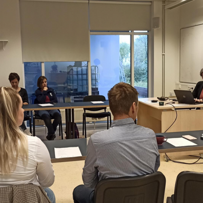 Zdjęcie przedstawia grupę osób siedzących przy stołach na spotkaniu edukacyjnym oraz osobę prowadzącą , która siedzi przy biurku przed laptopem oraz drugą osobę prowadzącą, która stoi przy tablicy.