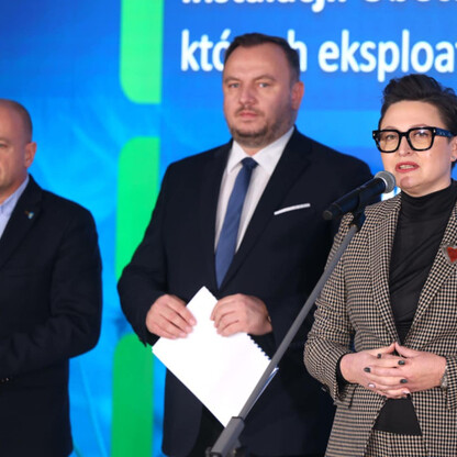 Podczas konferencji Marszałkowskiego Programu Poprawy Jakości Powietrza w Śląskim, jedna z uczestniczek przemawia do mikrofonu, podczas gdy dwaj inni słuchacze pozostają w tyle. (wygenerowano przez AI)