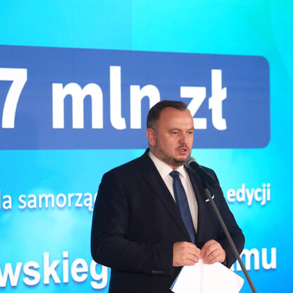 Mężczyzna w garniturze przemawia na podium, podczas konferencji prasowej dotyczącej finansowania programu poprawy jakości powietrza w Śląsku. (wygenerowano przez AI)
