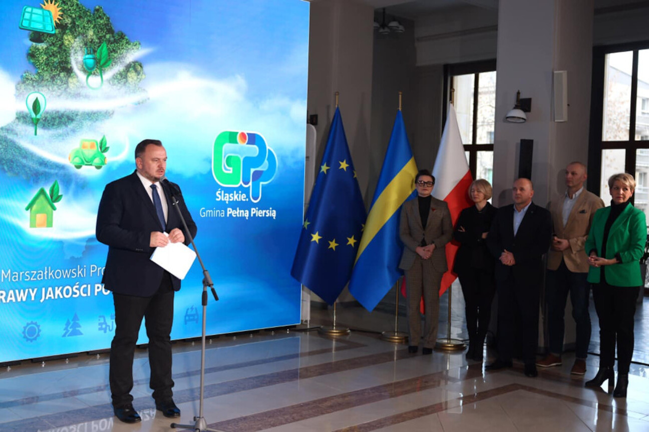 Podczas konferencji dotyczącej poprawy jakości powietrza województwa śląskiego, dwie osoby stoją na podium, jedna przemawia do mikrofonu, druga trzyma dokument. (wygenerowano przez AI)