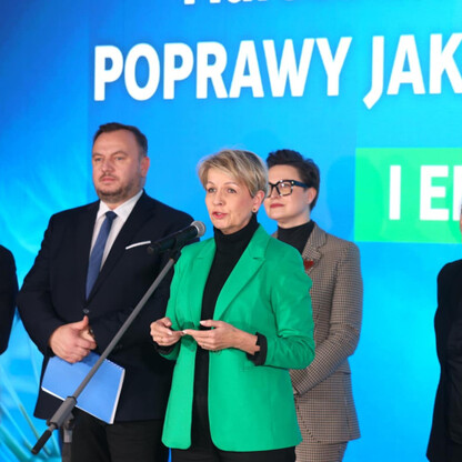 Grupa osób w garniturach i zielonym płaszczu stoi przed ekranem z napisem 