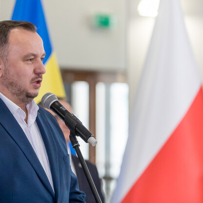 Mężczyzna w niebieskim garniturze przemawia na konferencji prasowej, przed mikrofonami i flagami UE oraz polskimi. (wygenerowano przez AI)