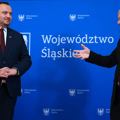 Dwóch mężczyzn w strojach formalnych trzyma się za ręce przed emblematem i napisem 