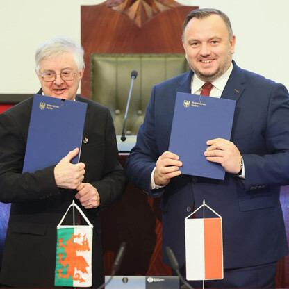 Dwóch mężczyzn w formalnym stroju, trzymających niebieskie foldery, stojących w sali parlamentarnej, podczas ceremonii podpisania Memorandum o współpracy między Województwem Śląskim a Rządem Walii. (wygenerowano przez AI)