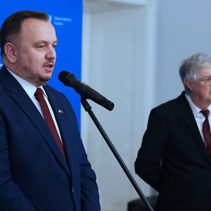Dwa mężczyźni w garniturach mówią na mikrofonach przed niebieskim tłem z napisem 