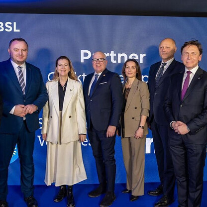 Grupa siedmiu ludzi w strojach formalnych stoi przed tłem z logo i nazwą 