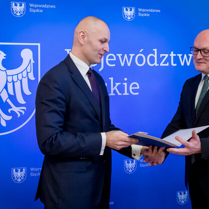 Dwa mężczyźni w strojach urzędniczych wręczają dokument, przedstawiając projekt koordynacji i monitorowania edukacji województwa śląskiego. (wygenerowano przez AI)