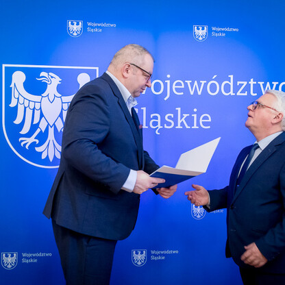 Dwóch mężczyzn w strojach formalnych trzyma się za ręce przed emblematem Województwa Śląskiego. (wygenerowano przez AI)