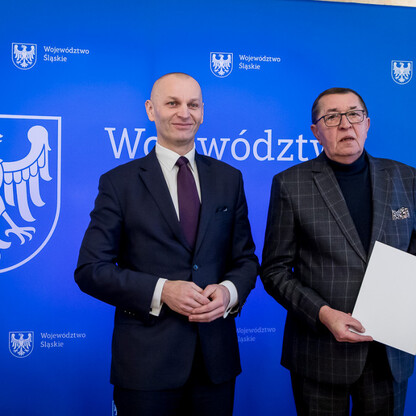Dwóch mężczyzn w garniturach przedstawiających Silesia, jednego z nich w ciemnym garniturze trzymającego dokument, drugiego w karo, na tle niebieskiego plakatu z emblematem i napisami Silesia. (wygenerowano przez AI)