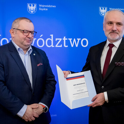 Dwóch mężczyzn w strojach formalnych stoi przed niebieskim tłem z emblematem Województwa Śląskiego, jednego wręcza dokument, drugi trzyma mu rękę. (wygenerowano przez AI)