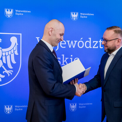 Dwóch mężczyzn w strojach formalnych trzyma się za ręce przed emblematem i napisem 