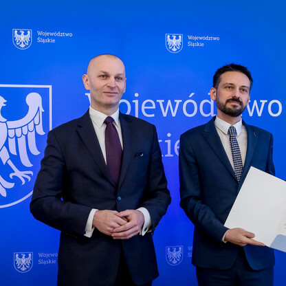 Dwóch mężczyzn w strojach urzędniczych stoi przed niebieskim tłem z herbem Województwa Śląskiego, jednym trzyma dokument, drugi ma ręce skrzyżowane. (wygenerowano przez AI)
