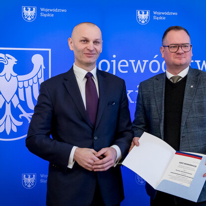 Dwóch mężczyzn w formalnym stroju stoi przed niebieskim tłem z herbem Województwa Śląskiego, jednym z nich trzyma dokument 
