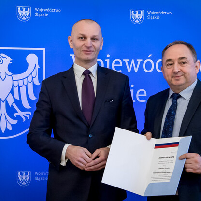 Dwóch mężczyzn w strojach formalnych stoi przed niebieskim tłem z herbem Województwa Śląskiego, trzymając dokument 