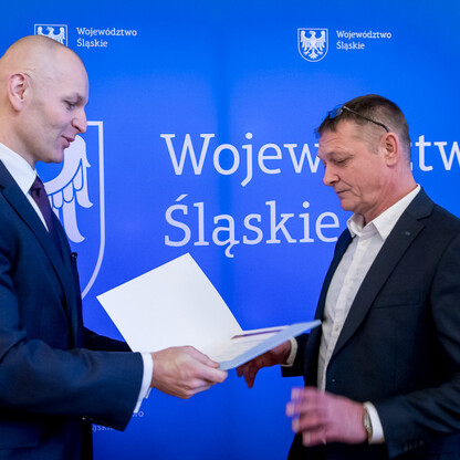 Dwóch mężczyzn w strojach formalnych przekazujących dokument, symbolizujący współpracę w projekcie koordynacji i monitorowania edukacji województwa śląskiego. (wygenerowano przez AI)