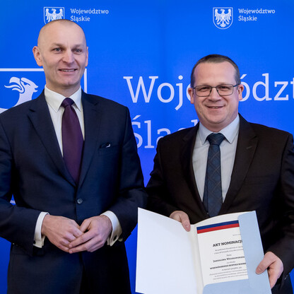 Dwóch mężczyzn w strojach formalnych stoi przed niebieskim tłem z emblematem i napisem 