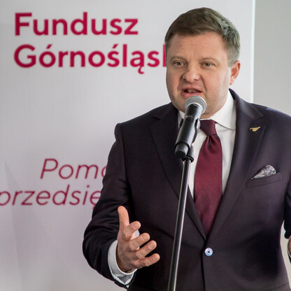 Mężczyzna w garniturze przemawia na podium z mikrofonem, tło z napisem 