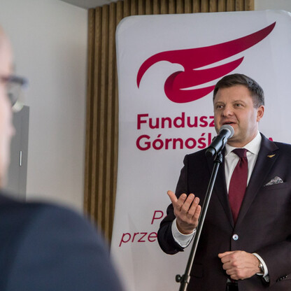 Mężczyzna w garniturze przemawia do mikrofonu na podium, promując działania Funduszu Górnośląskiego na rzecz rozwoju eksportu dla małych i średnich przedsiębiorstw w Śląsku. (wygenerowano przez AI)