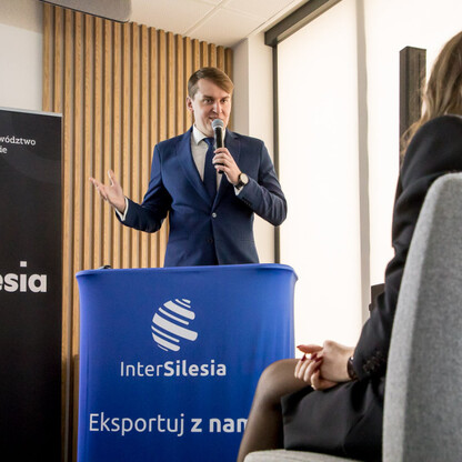 Mężczyzna w garniturze przemawia na podium z mikrofonem, wspierając rozwój eksportu województwa śląskiego. (wygenerowano przez AI)