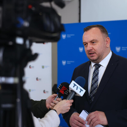 Mężczyzna w garniturze odpowiada na pytania dziennikarzy podczas konferencji prasowej, dotyczącej budowy mostu na Odrze. (wygenerowano przez AI)