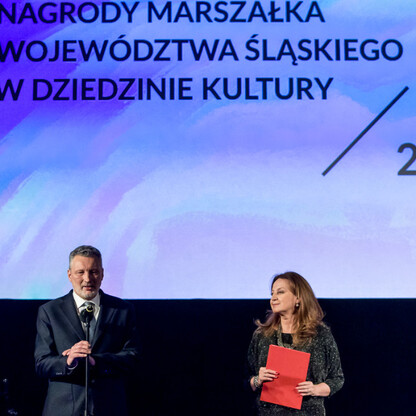 Podczas ceremonii wręczenia nagród Marszałka Województwa Śląskiego w dziedzinie kultury, trzech osób stoi na scenie, jedna trzyma certyfikat, druga mówi. (wygenerowano przez AI)