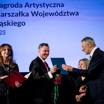 Grupa ludzi uczestniczy w ceremonii wręczenia nagród artystycznych w Katowicach, podczas której jedna osoba odbiera certyfikat. (wygenerowano przez AI)