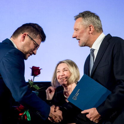 Podczas uroczystości wręczenia nagród Marszałka Województwa Śląskiego Culture Awards, dwaj mężczyźni uściskają ręce, jednym z nich trzymając certyfikat i różę, symbolizujące uznanie za wkład w kulturę (wygenerowano przez AI)