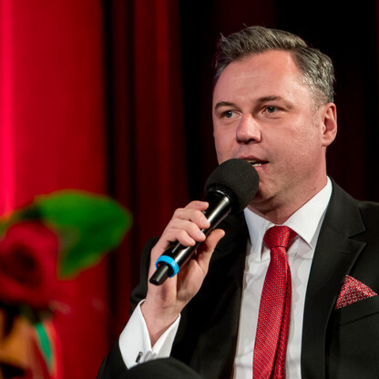 Mężczyzna w garniturze i czerwonym krawacie przemawia do mikrofonu w czerwonym teatrze, podczas ceremonii wręczenia nagród kulturalnych w Silesia. (wygenerowano przez AI)