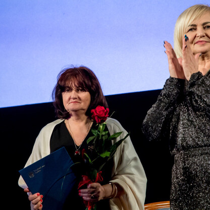 Dwie kobiety na scenie podczas uroczystości wręczenia nagród Marszałka Województwa Śląskiego Culture Awards. Jedna trzyma bukiet i certyfikat, druga klapsuje. Atmosfera formalna i celebracyjna. (wygenerowano przez AI)