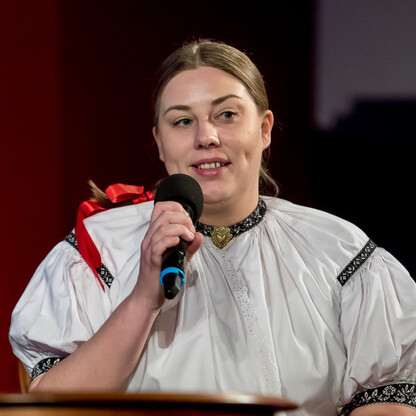 Podczas uroczystości wręczenia nagród Marszałkowskich w Silesia, osoba ubrana w tradycyjną białą bluzę z czarnymi i czerwonymi akcentami, trzymająca mikrofon, wygłasza przemówienie na scenie podczas c (wygenerowano przez AI)
